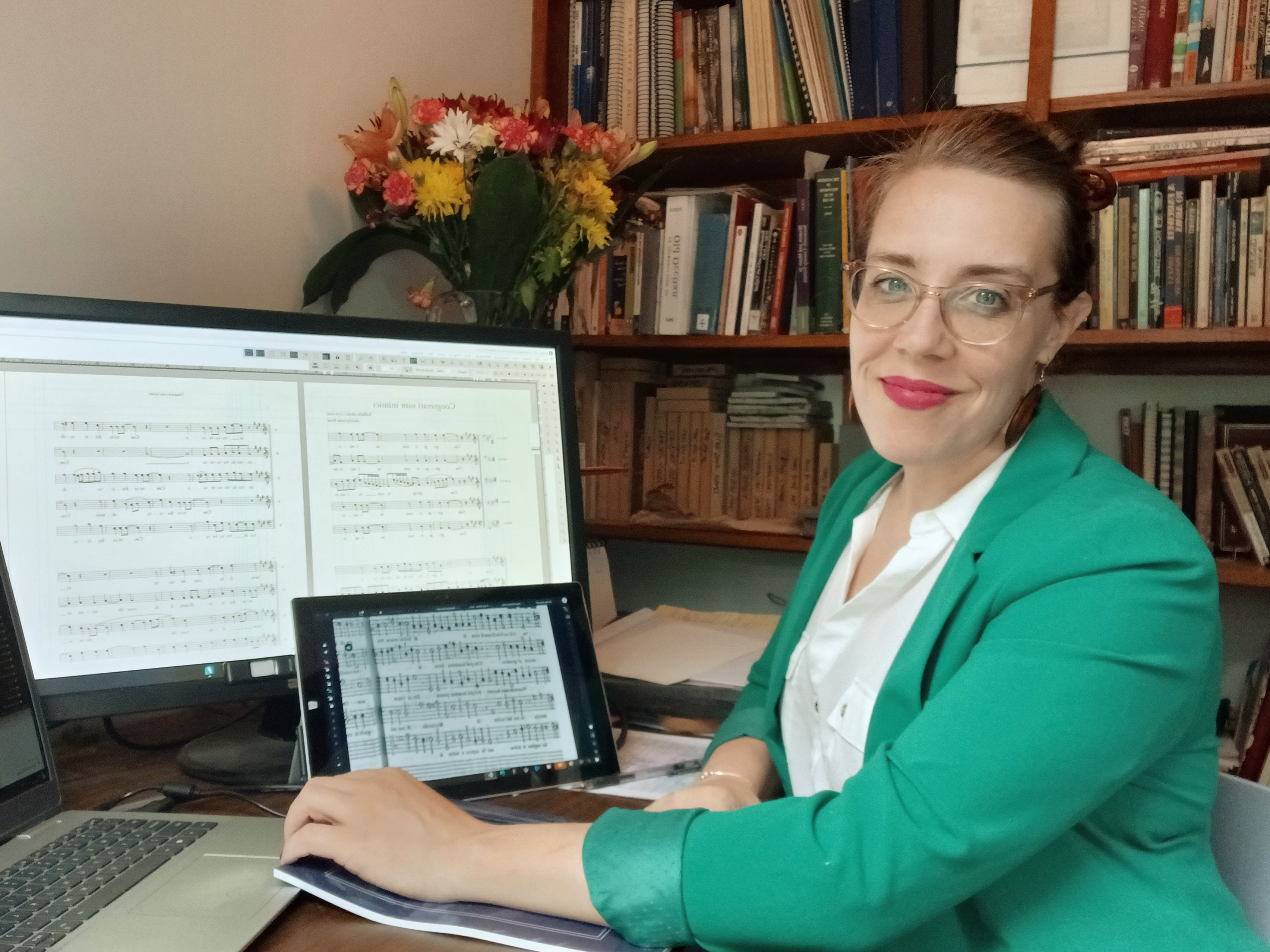 photo of a smiling woman wearing a green blazer and sitting at a desk with music notation software on her laptop screen.